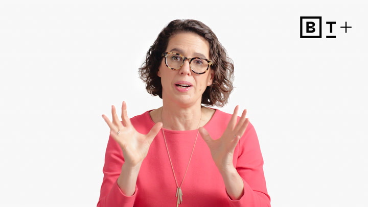A woman with curly brown hair and glasses, wearing a pink top and pendant necklace, gestures with both hands in front of a plain white background with a "B T +" logo.