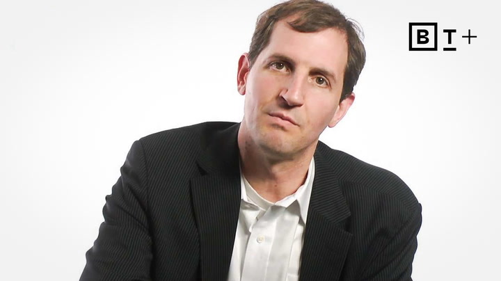 A man in a dark suit and white shirt sits against a plain white background with a "B T +" logo in the top right corner.