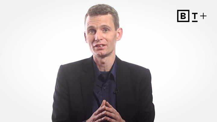 A man in a dark suit and shirt sits against a plain white background, hands clasped, with a "BT+" logo in the top right corner.