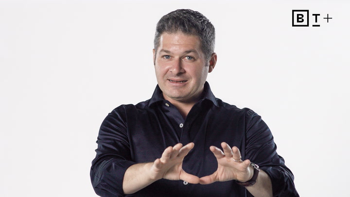 A man in a dark shirt gestures with his hands in front of a white background. The letters "BT+" appear in the top right corner.