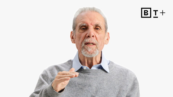 An older man with gray hair and beard, wearing a light blue collared shirt and gray sweater, gestures with one hand. A "B T +" logo is visible in the top right corner.