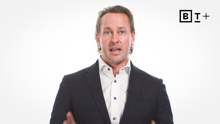 A man in a dark suit and white shirt speaks in front of a white background with the B+T Plus logo in the top right corner.