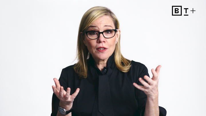 A woman with blonde hair and glasses, dressed in black, gestures with her hands while speaking against a plain white background with a "BT+" logo in the top right corner.
