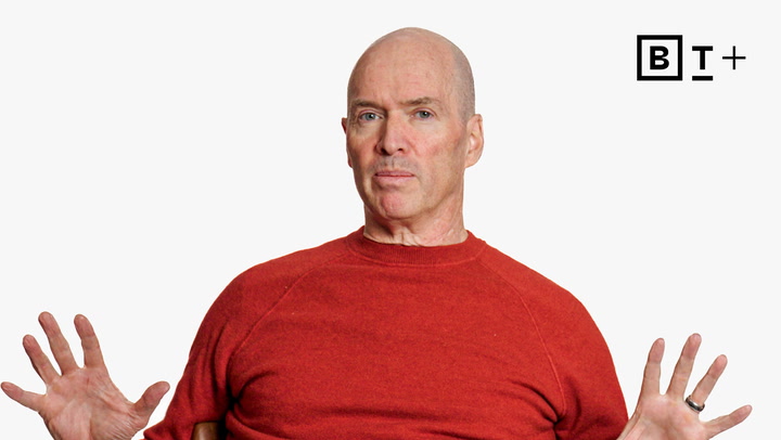 A bald man in a red sweater sits with hands raised at chest level against a plain white background with a "BT+" logo in the top right corner.