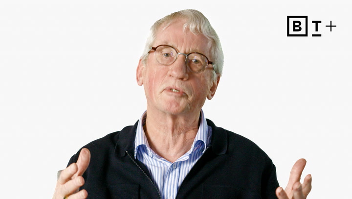 An older man with glasses and gray hair speaks while gesturing with his hands. He is wearing a dark jacket over a striped shirt. BT+ logo is visible in the top right corner.