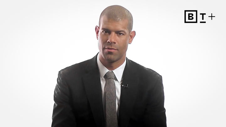 A man in a dark suit and tie sits against a plain white background with a "B T +" logo in the top right corner.