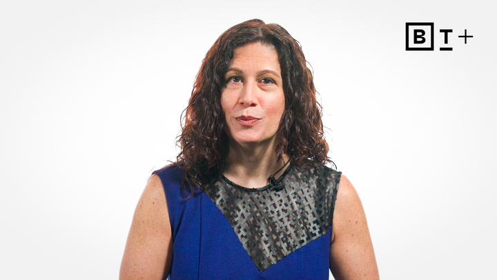 A woman with curly brown hair wearing a blue sleeveless top stands against a white background with the letters "BT+" in the top right corner.