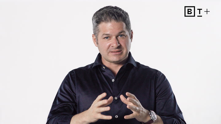 A man in a dark shirt speaks while gesturing with his hands in front of a plain white background with a "BT+" logo in the top right corner.