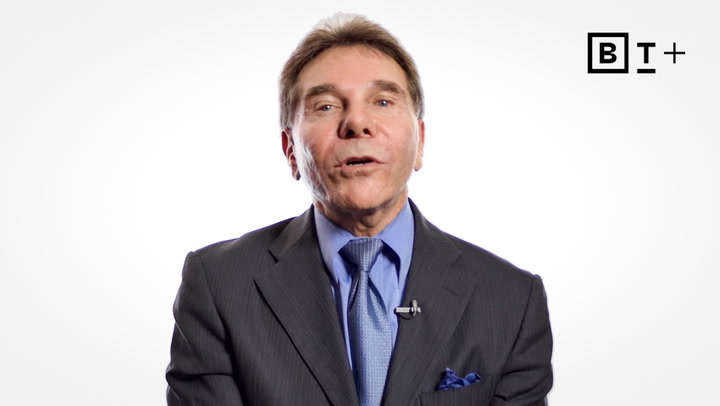 A man in a blue suit and tie speaks in front of a plain white background with a microphone attached to his lapel and the B+ logo in the top right corner.