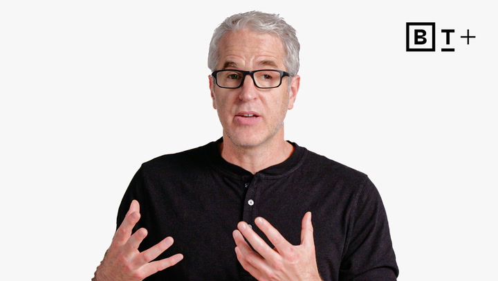 A man with gray hair and glasses, wearing a black shirt, gestures with his hands while speaking in front of a plain white background with a "BT+" logo in the corner.