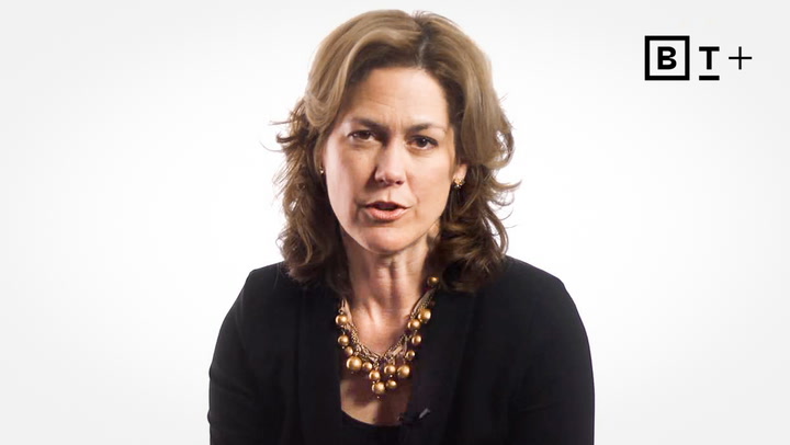 A woman with wavy brown hair, wearing a black top and a chunky gold necklace, sits against a plain white background with a "B T +" logo in the upper right corner.