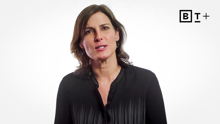 A woman with shoulder-length brown hair wearing a black blouse stands against a white background with a "B T +" logo in the top right corner.