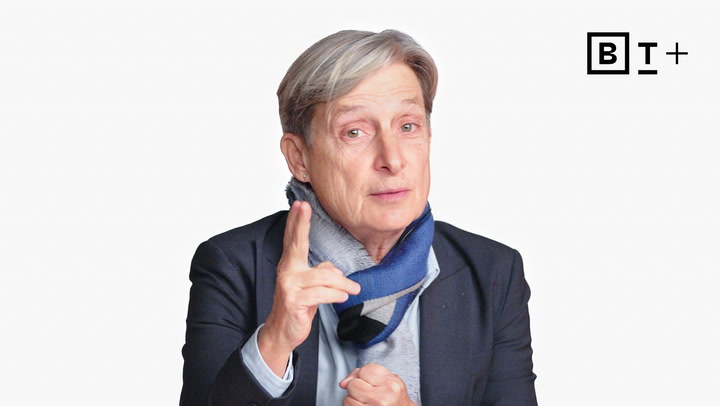 A person with short gray hair wearing a suit jacket and blue scarf gestures with one hand. The letters "BT+" appear in the top right corner.