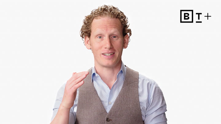A man with curly hair wearing a light blue shirt and gray vest gestures with his right hand. The background is white with a "B T +" logo in the top right corner.