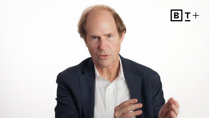 A middle-aged man in a suit gestures with his hands while speaking against a plain white background with a "BT+" logo in the top right corner.