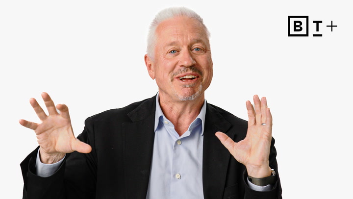 An older man with white hair and a beard, wearing a black blazer and light blue shirt, gestures with both hands while speaking. The "B T +" logo appears in the top right corner.