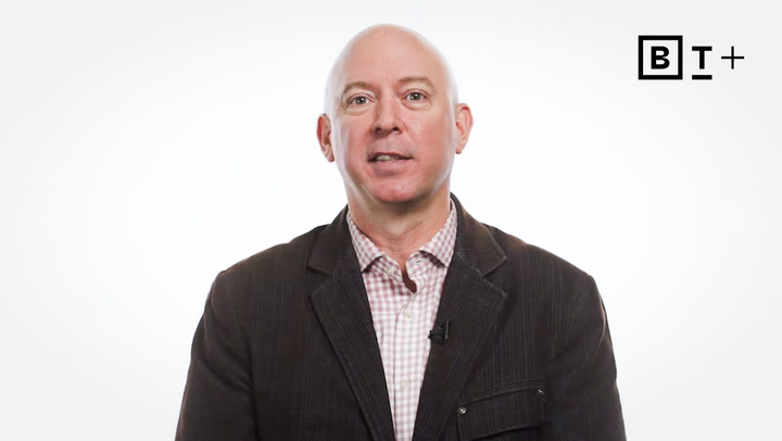 A bald man in a dark jacket and checkered shirt stands against a plain white background with a "B T +" logo in the top right corner.