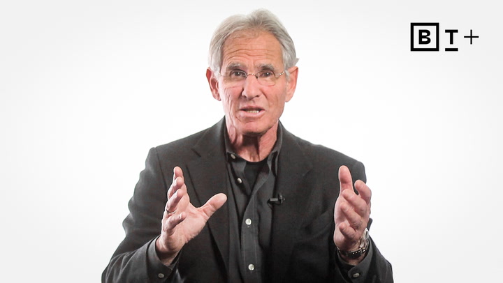An older man with gray hair and glasses, wearing a dark suit, speaks while gesturing with both hands against a plain white background with a "BT+" logo in the corner.