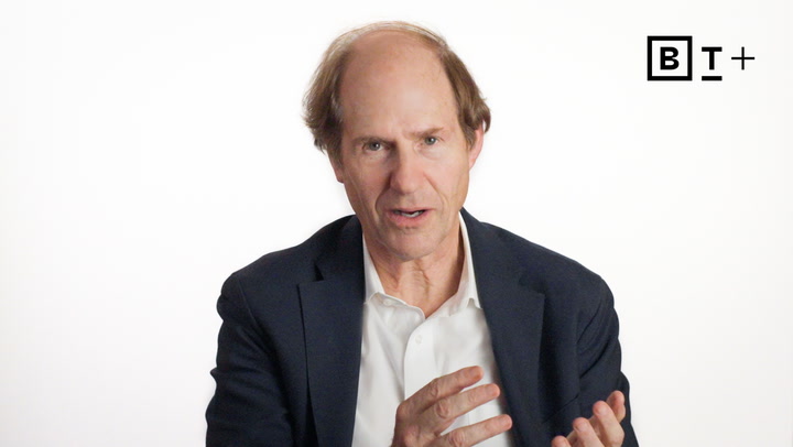 A middle-aged man in a suit speaks while gesturing with his hands against a plain white background with a "B T +" logo in the top right corner.