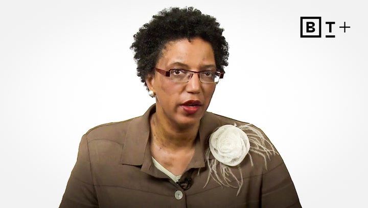 A person with short curly hair and glasses wears a brown jacket with a white flower brooch, sitting against a plain white background with a "BT+" logo in the corner.