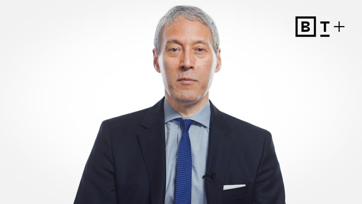 A man with gray hair in a dark suit, blue shirt, and blue tie sits against a white background with the letters "BT+" in the top right corner.