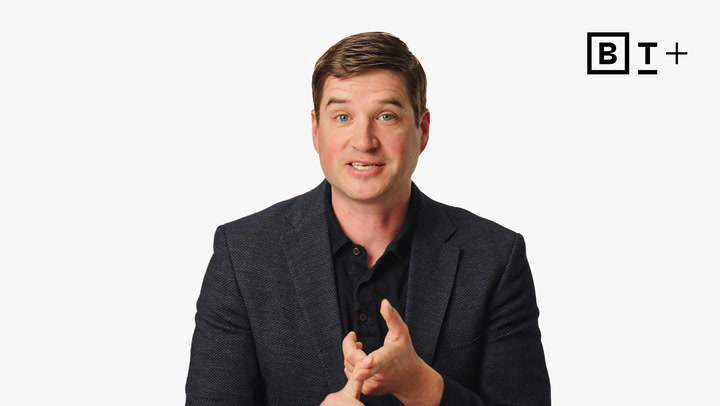 A man in a dark blazer speaks while gesturing with his hands. There is a "B T +" logo in the top right corner of a plain white background.