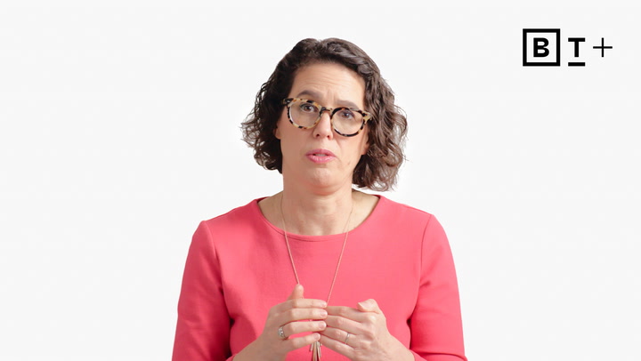 A woman with short curly hair and glasses, wearing a coral top, gestures with her hands while speaking in front of a plain white background with a "B T +" logo.
