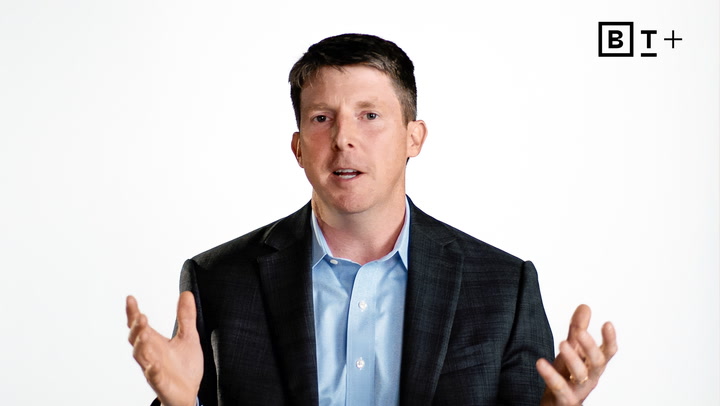 A man in a dark suit jacket and blue shirt speaks with his hands raised, against a plain white background with a "B T +" logo in the top right corner.