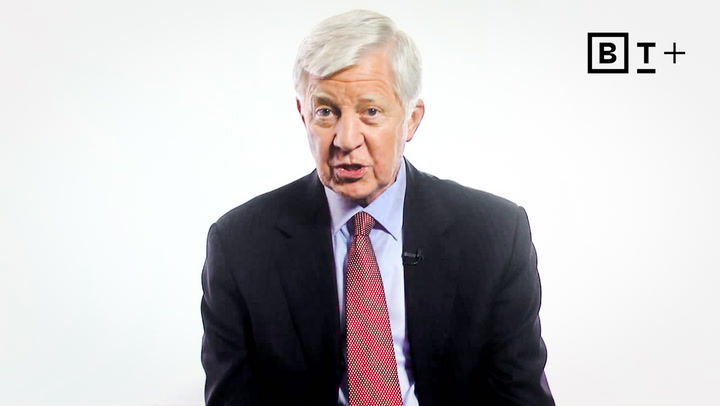 An older man in a dark suit and red tie speaks while seated against a plain white background with a "BT+" logo in the top right corner.