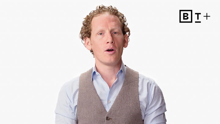 A man with curly hair wearing a light blue shirt and brown vest speaks in front of a plain white background with the B+T logo in the top right corner.