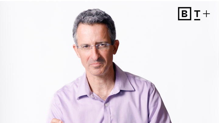A man with short gray hair and glasses wearing a light purple shirt stands against a white background with a black "BT+" logo in the top right corner.
