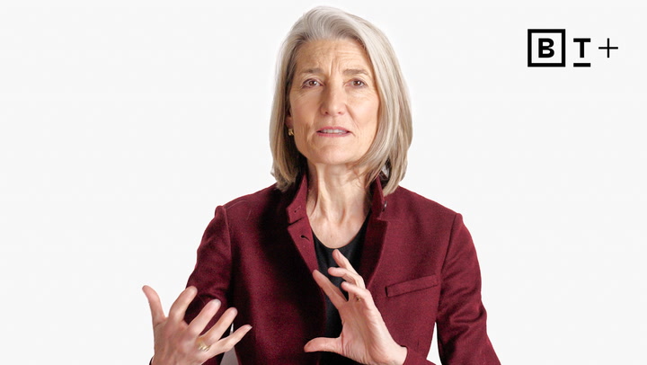 A woman with gray hair wearing a burgundy blazer gestures with her hands while speaking, with the letters "B T +" visible in the top right corner.