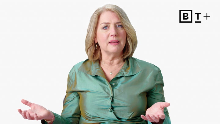 A woman with blonde hair in a green satin blouse gestures with open hands while speaking, in front of a plain white background with a "B T +" logo in the top right corner.