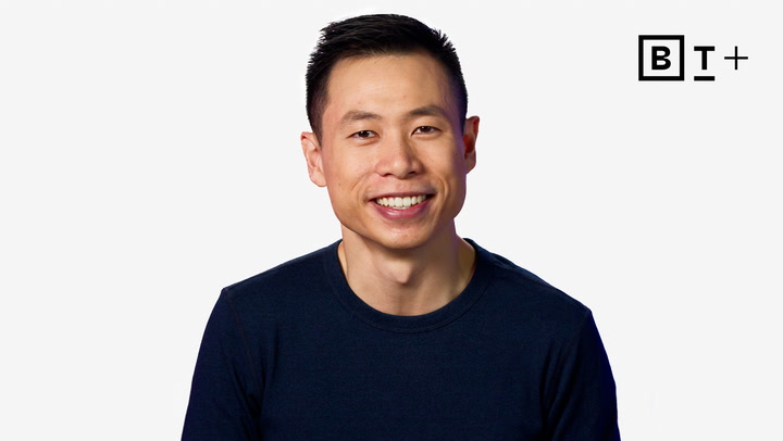 A man in a dark shirt smiles at the camera in front of a plain white background with a "B T +" logo in the top right corner.