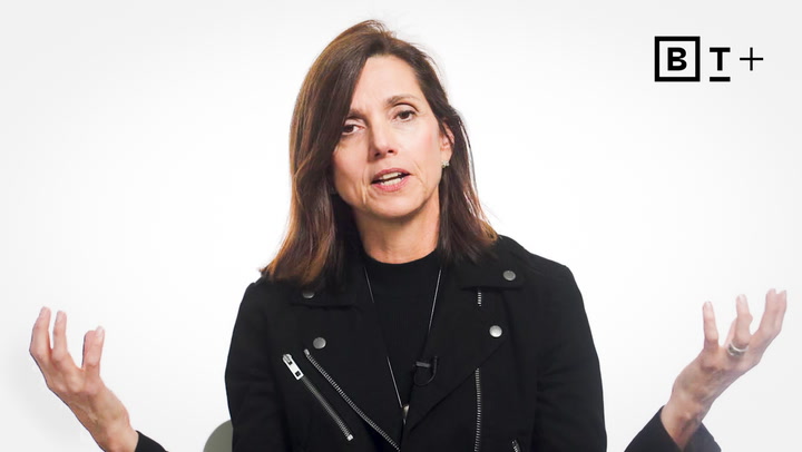 A woman with shoulder-length brown hair wearing a black jacket gestures with both hands while speaking. A "BT+" logo appears in the top right corner.