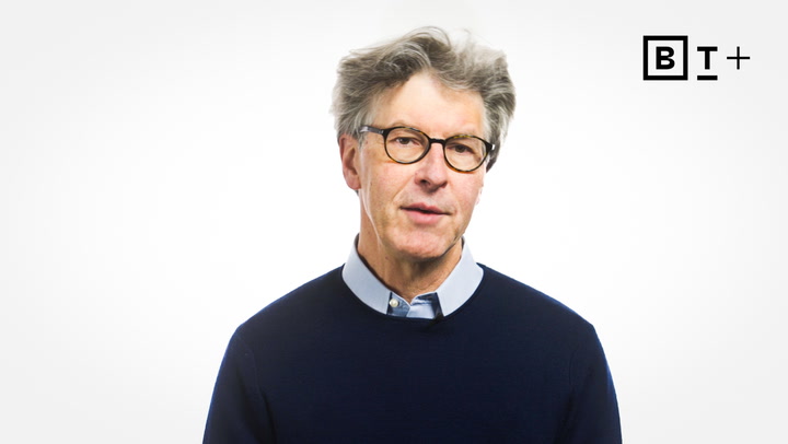 An older man with gray hair, glasses, and a navy sweater over a collared shirt stands in front of a plain white background with a "B T +" logo in the top right corner.