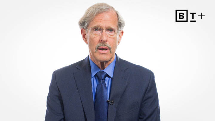 An older man with gray hair, glasses, and a mustache wears a blue suit and tie, speaking in front of a plain white background with a "B T +" logo in the corner.