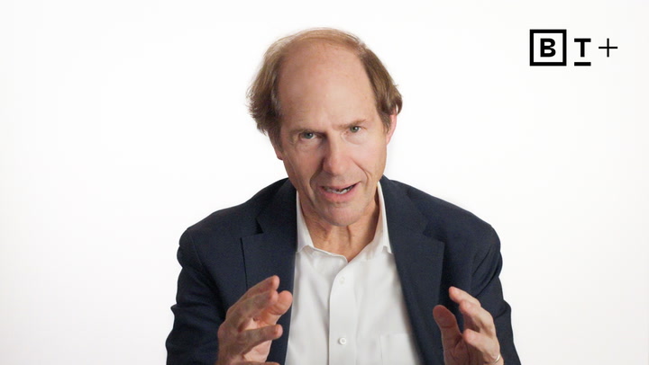 A middle-aged man in a suit gestures with his hands while speaking in front of a plain white background with a BT+ logo in the top right corner.
