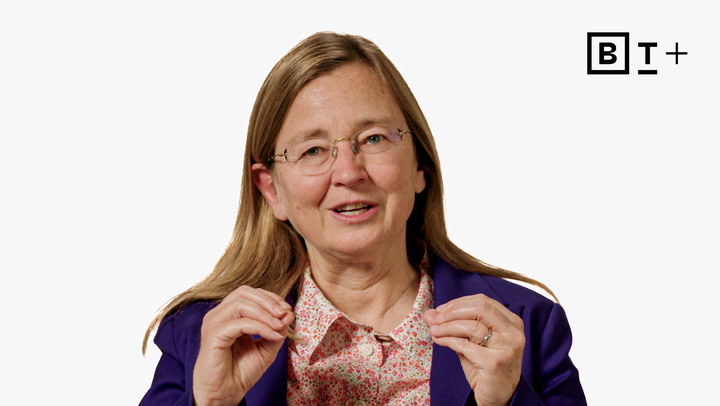 A woman with long hair and glasses, wearing a purple blazer and patterned shirt, gestures with her hands while speaking, with a black "B T +" logo in the top right corner.