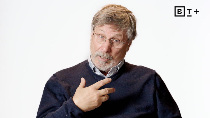 An older man with glasses and a beard, wearing a dark sweater over a collared shirt, gestures with his hand while speaking against a plain white background. BT+ logo in top right corner.