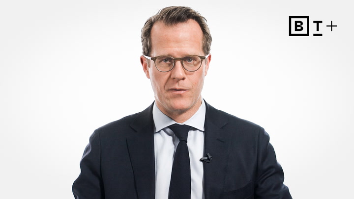 A man wearing glasses and a suit sits in front of a white background with a small BT+ logo in the top right corner.