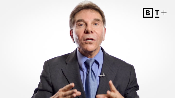 A man in a suit and tie speaks, gesturing with his hands in front of a plain white background with a "BT+" logo in the top right corner.