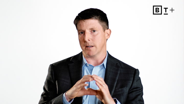 A man in a dark suit and light blue shirt speaks, gesturing with his hands against a plain white background with a "BT+" logo in the top right corner.