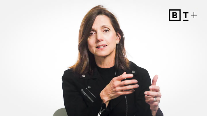 A woman with shoulder-length brown hair, wearing a black jacket, gestures while speaking in front of a plain white background with a "BT+" logo in the corner.