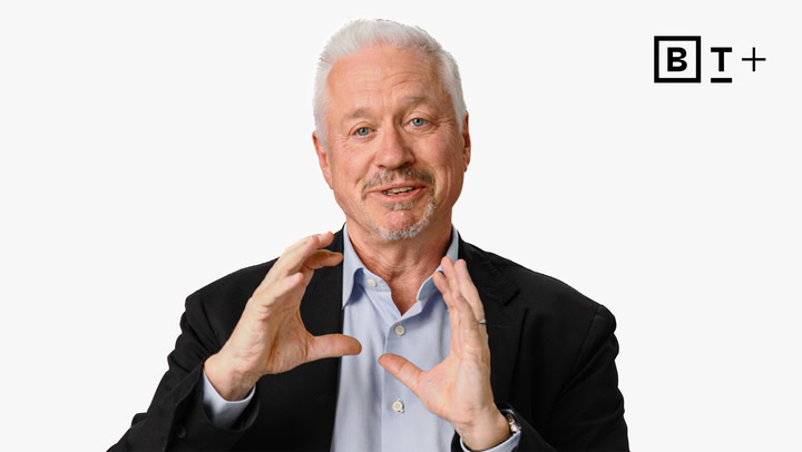 A man with white hair and a goatee in a suit gestures with his hands while speaking, with a "B T +" logo in the top right corner on a plain background.