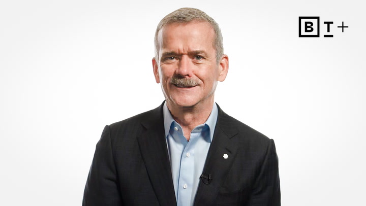 A man in a dark suit jacket and light blue shirt is smiling in front of a plain white background with a "B T +" logo in the upper right corner.
