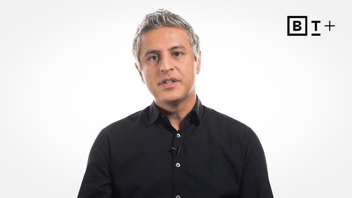 A man with gray hair wearing a black button-up shirt sits in front of a plain white background with a "B T +" logo in the top right corner.