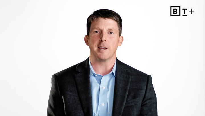 A man in a dark suit and blue shirt speaks in front of a plain white background with a "B T +" logo in the top right corner.