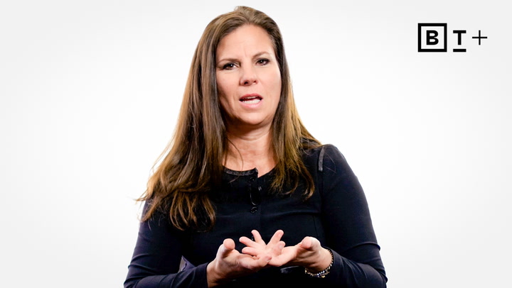 A woman with long brown hair in a black top speaks and gestures with her hands in front of a white background with a "B T +" logo in the corner.