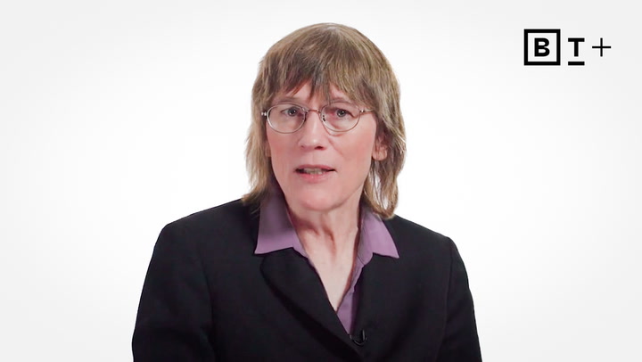 A person with shoulder-length hair and glasses, dressed in a suit jacket and collared shirt, sits in front of a plain white background with a BT+ logo in the upper right corner.